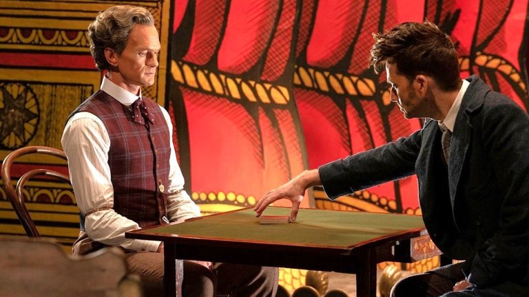 Il Toymaker di Neil Patrick Harris e il Dottore di David Tennant hanno un teso faccia a faccia su un tavolo di fronte a un palco in Doctor Who: The Giggle. 