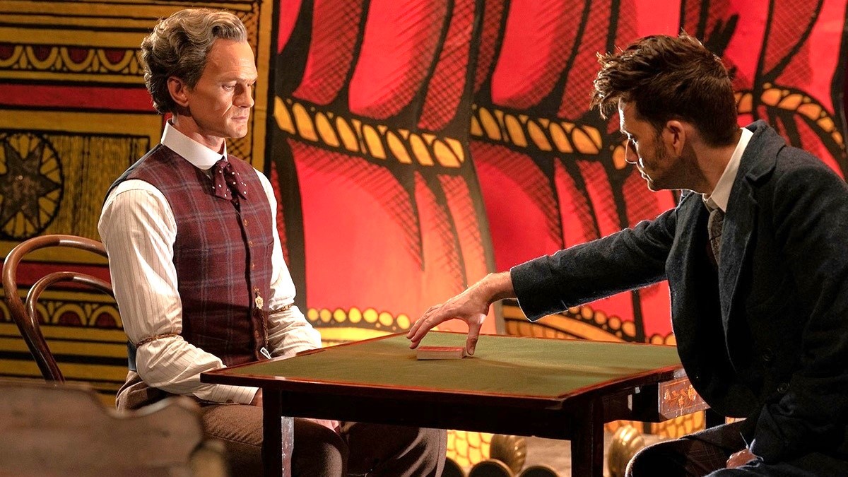 Il Toymaker di Neil Patrick Harris e il Dottore di David Tennant hanno un teso faccia a faccia su un tavolo di fronte a un palco in Doctor Who: The Giggle.
